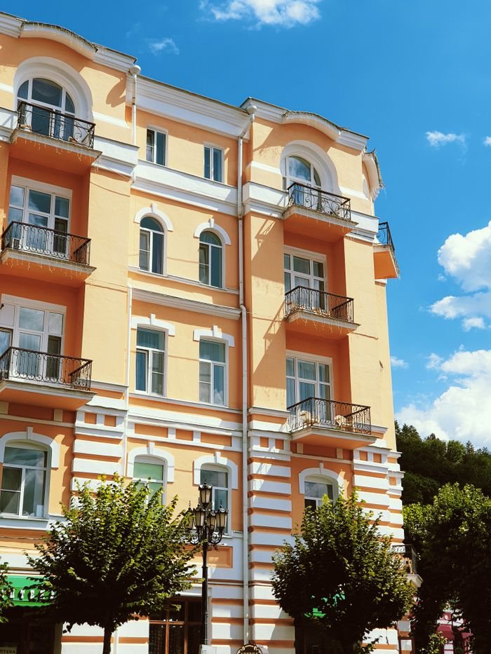 our-story-01 Charming yellow residential building with balconies and trees, set against a vibrant blue sky on a sunny day.